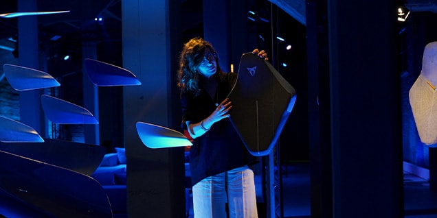 A woman with brown hair is holding a dark, shield-shaped object with a copper-colored Cupra logo on it. 