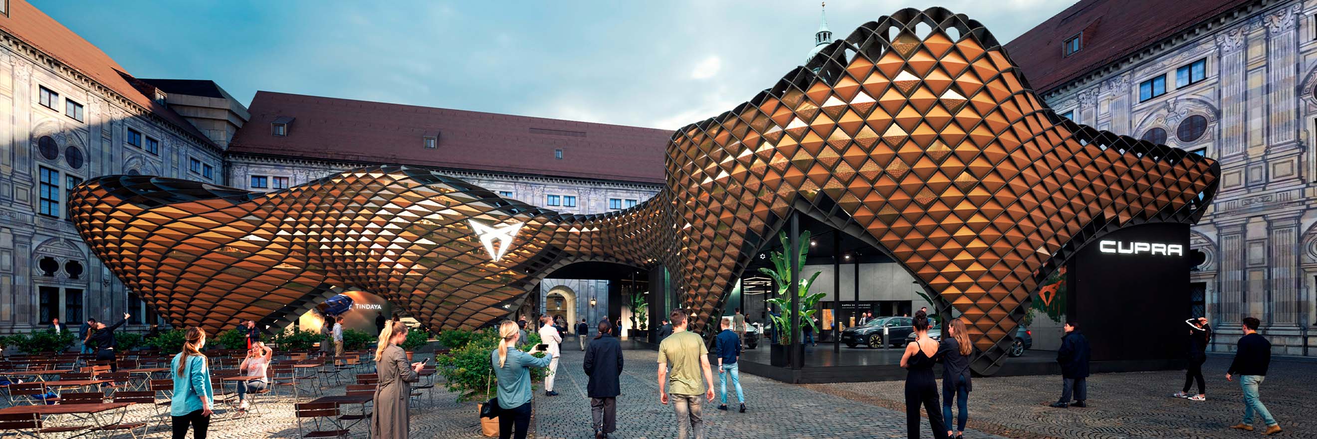 A modern, undulating architectural structure with a pattern of interconnected, metallic diamond shapes dominates the center of the image. The structure is an outdoor pavilion with a large opening in the middle, revealing a Cupra brand logo and a glimpse of an interior space. 