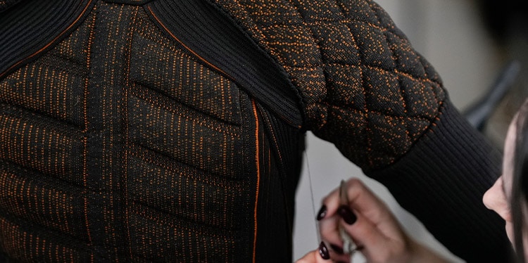 A close-up shot of the back of a black jacket or garment with intricate orange woven patterns. The garment has a quilted texture on the upper back and shoulder, while the lower back features a ribbed pattern.