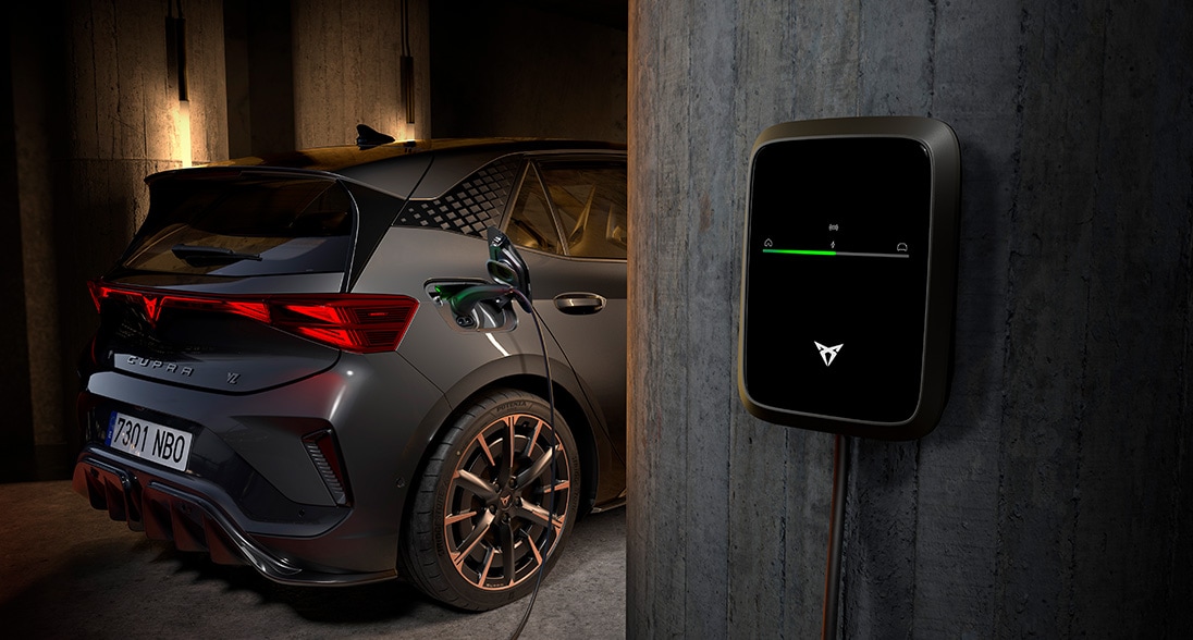 A dark grey Cupra electric vehicle parked in a dimly lit, modern concrete garage, plugged into a black, wall-mounted Cupra charging station that features a glowing green indicator light.