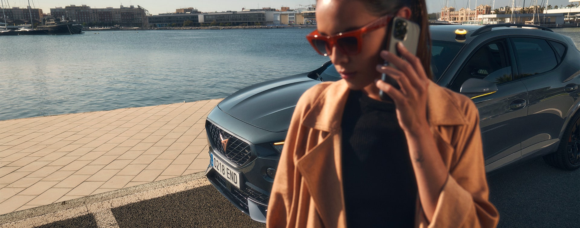 A woman wearing a tan trench coat and sunglasses talking on a smartphone in the foreground, with a CUPRA car parked behind her at a waterfront marina under a clear sky.