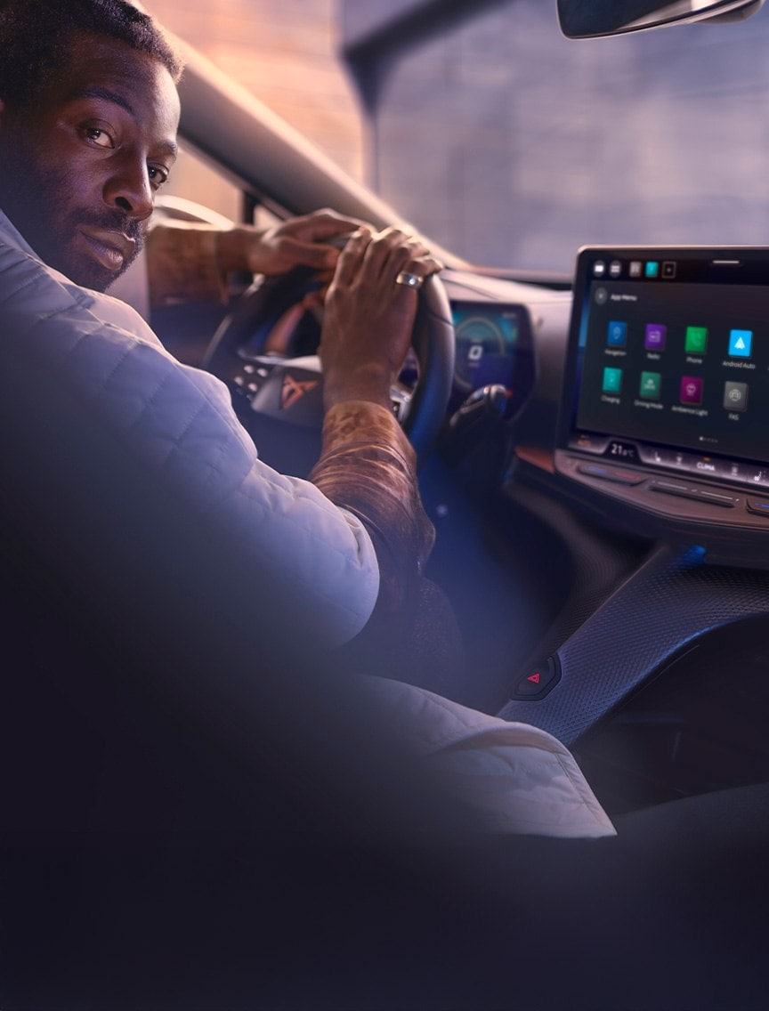 A man, wearing a white quilted jacket, is sitting in the driver's seat of a CUPRA Tavascan, turning to look over his shoulder. The car's steering wheel and a large central touchscreen display showing app icons are visible on the right.
