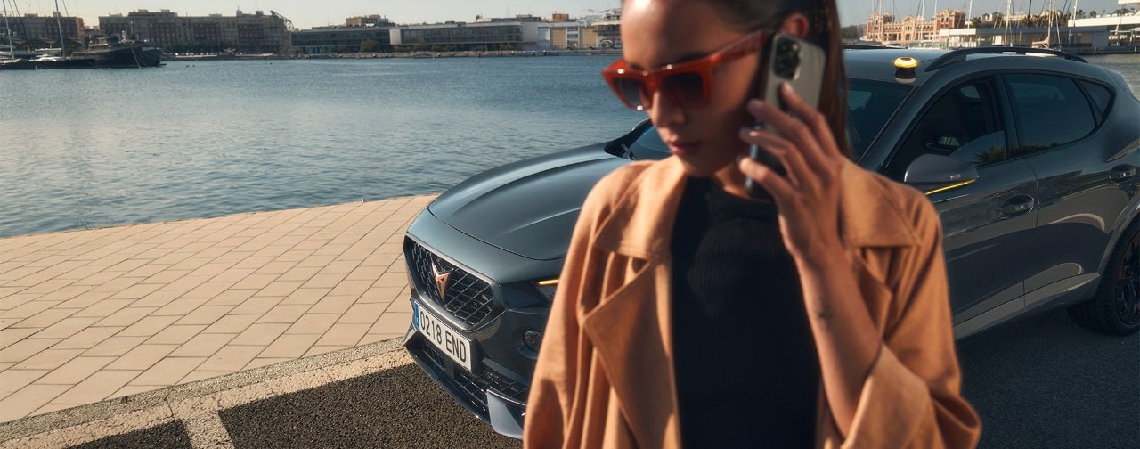 A woman wearing a tan trench coat and sunglasses talking on a smartphone in the foreground, with a CUPRA car parked behind her at a waterfront marina under a clear sky.