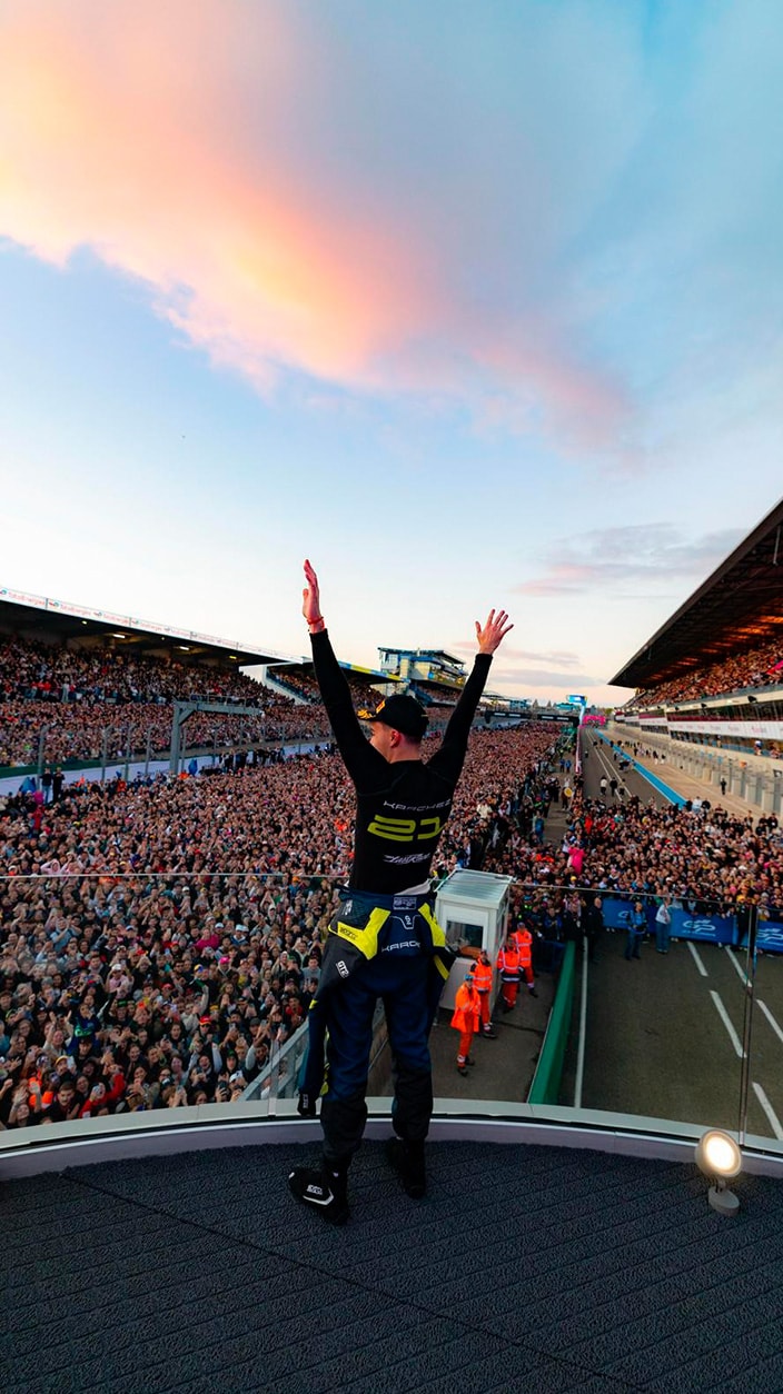 A victorious racing driver stands high on a podium, raising both arms in celebration towards a massive, cheering crowd filling the grandstands below. The scene unfolds at a major racetrack under a sky with pink and blue sunset clouds.