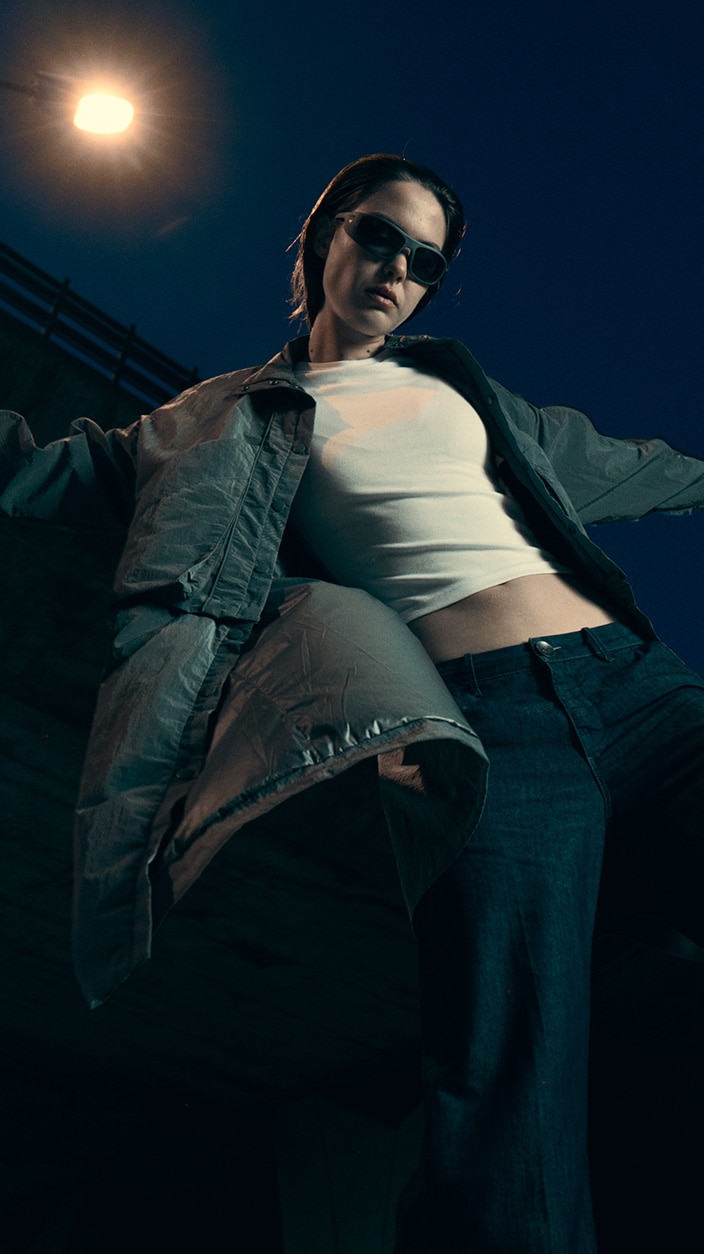 A dramatic, low-angle shot of a model showcasing the CUPRA collection, wearing dark sunglasses, a white crop top, dark trousers, and a voluminous open jacket. The urban night scene is illuminated by a glowing streetlamp against a dark blue sky. 