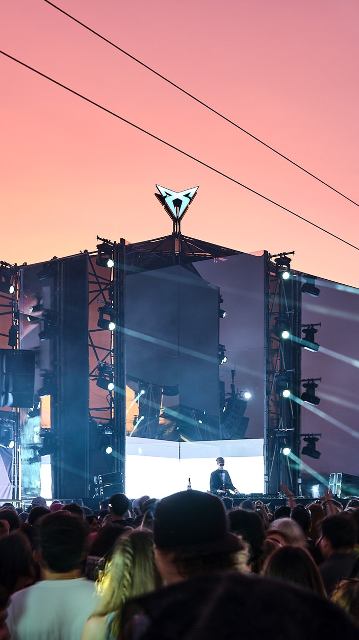 A vibrant outdoor music festival stage at sunset, featuring a DJ performing in front of a silhouetted crowd. The stage structure is illuminated by spotlights and topped with a prominently glowing CUPRA logo against a pink and orange sky.