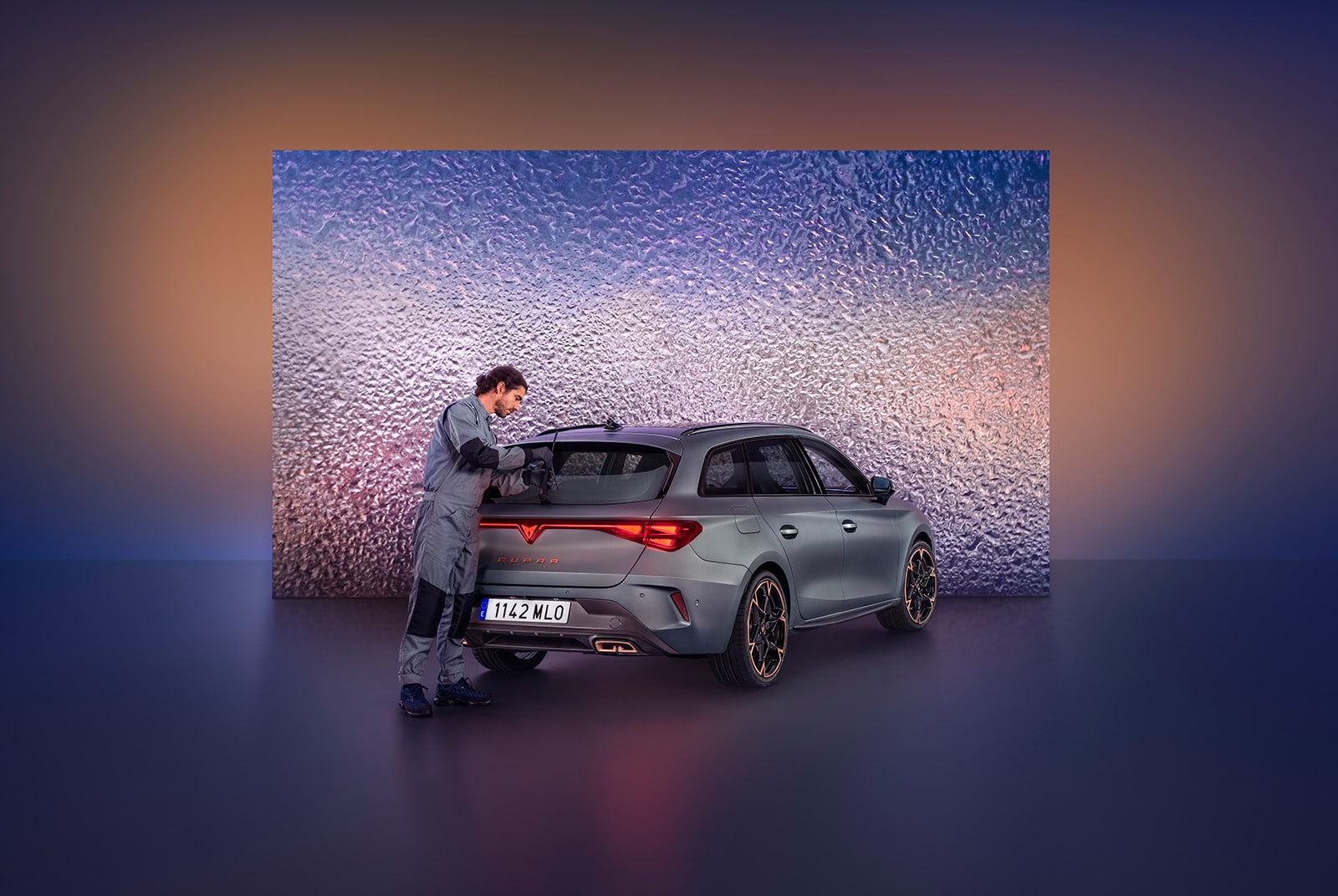A car technician in a grey jumpsuit carefully details the rear spoiler of a grey CUPRA car. The car is in a service bay, positioned in front of a large, illuminated wall with a textured surface that resembles rippling water.