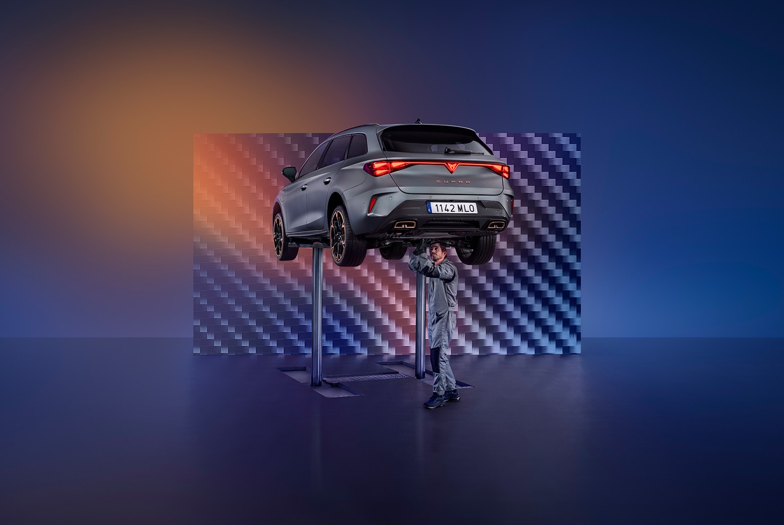 A car technician in a grey jumpsuit works on the undercarriage of a CUPRA car that is elevated high on a service lift. The scene is set in a modern garage with a large, patterned background and orange and blue lighting.