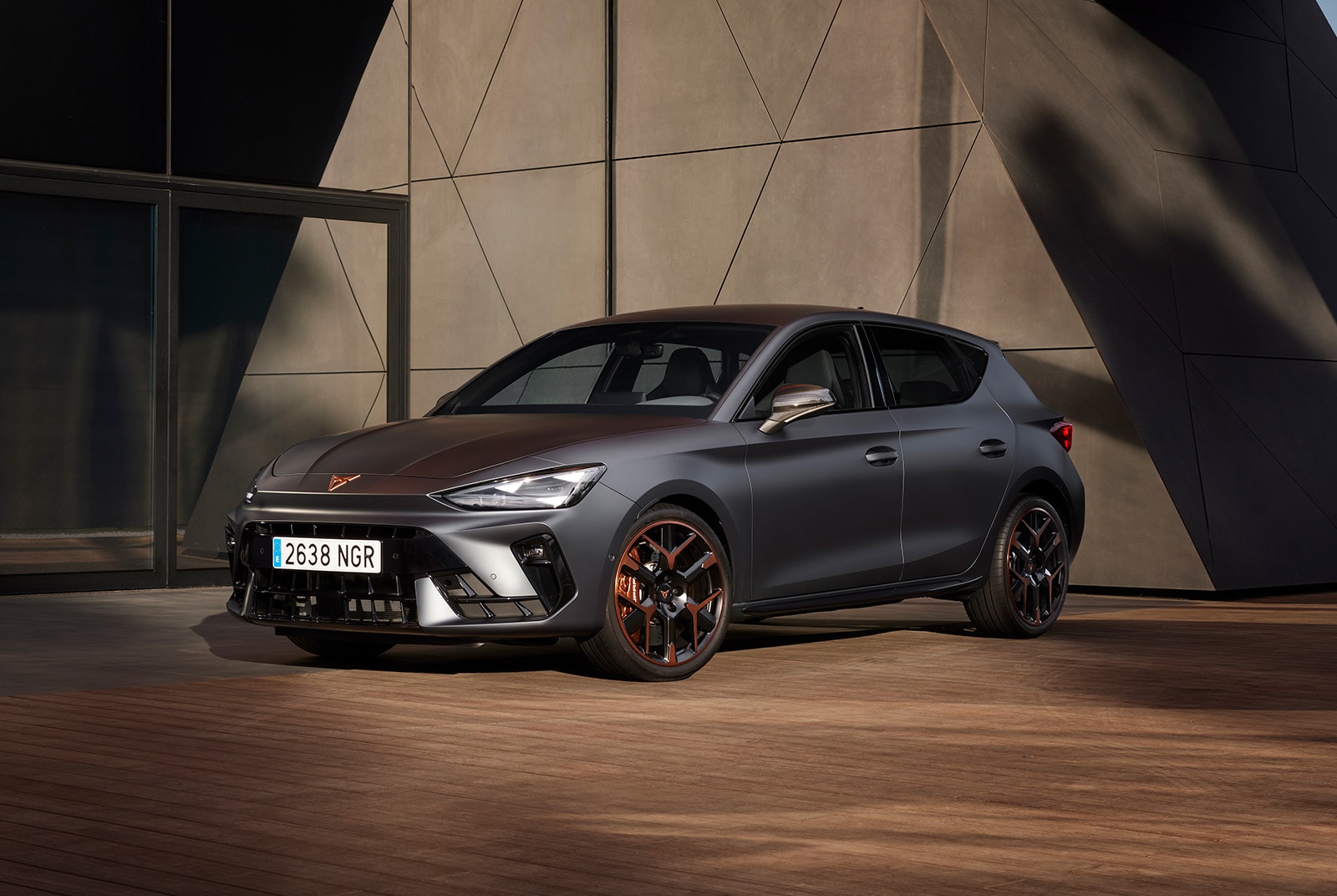 A CUPRA Leon parked on a wooden surface in front of a modern geometric building. The vehicle features black and copper alloy wheels and a copper emblem on the hood.