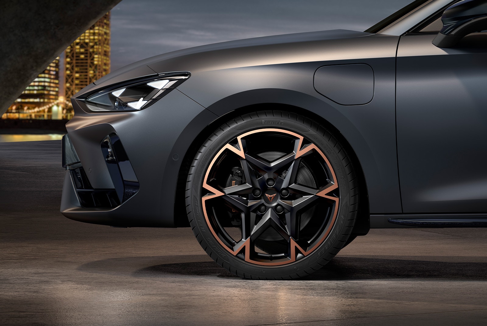 A close-up, low-angle shot of the front of a dark CUPRA car at night. The car's headlight is on, and the intricate black and bronze wheel is visible. In the blurred background, city lights are visible.