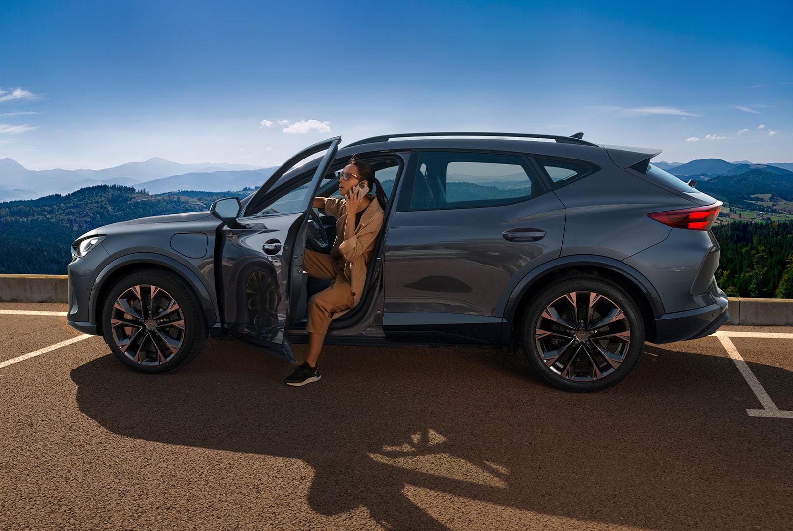 A person gets out of a dark gray CUPRA car, which is parked in a lot overlooking a scenic mountain range and forested hills. The person is holding a phone to their ear, with the car door open.