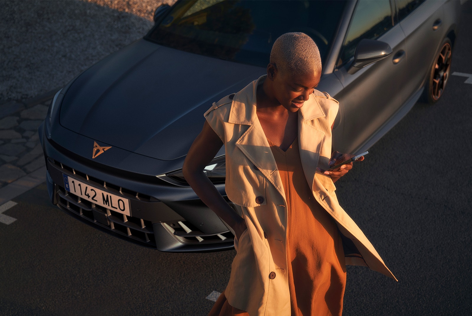 A person with short hair, wearing a brown dress and a light-colored trench vest, looks at their smartphone while walking away from a dark gray CUPRA car parked in a lot.