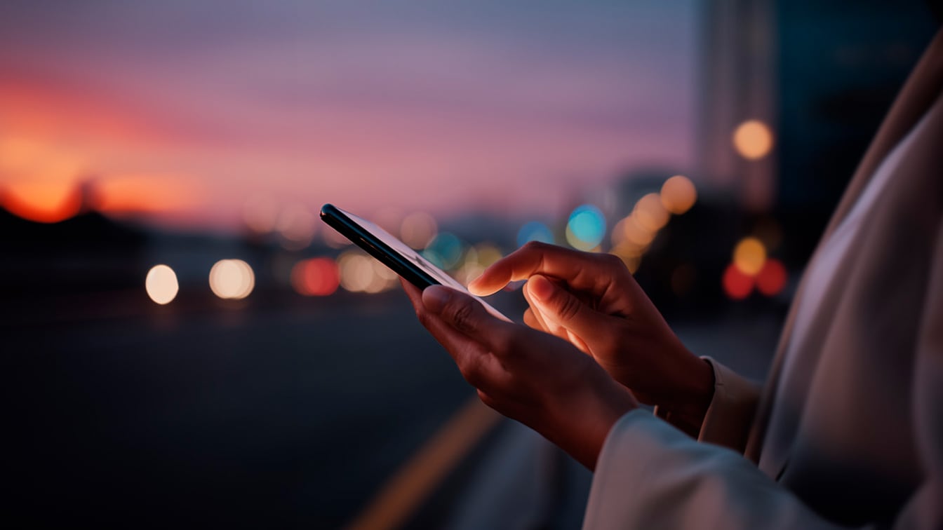 Close-up of hands using a glowing smartphone at dusk, set against a blurred city background with a vibrant orange and purple sunset and bokeh lights.