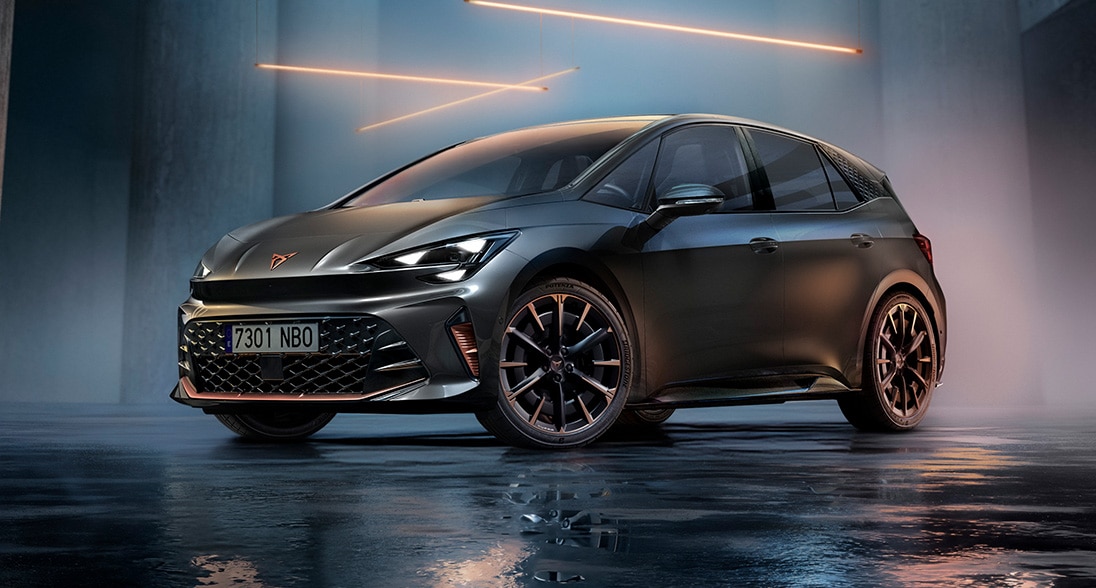 A CUPRA Born parked in a modern concrete architectural setting with a wet, highly reflective floor. The vehicle features illuminated sharp headlights and multi-spoke alloy wheels, highlighted by a series of glowing orange geometric light bars suspended overhead.