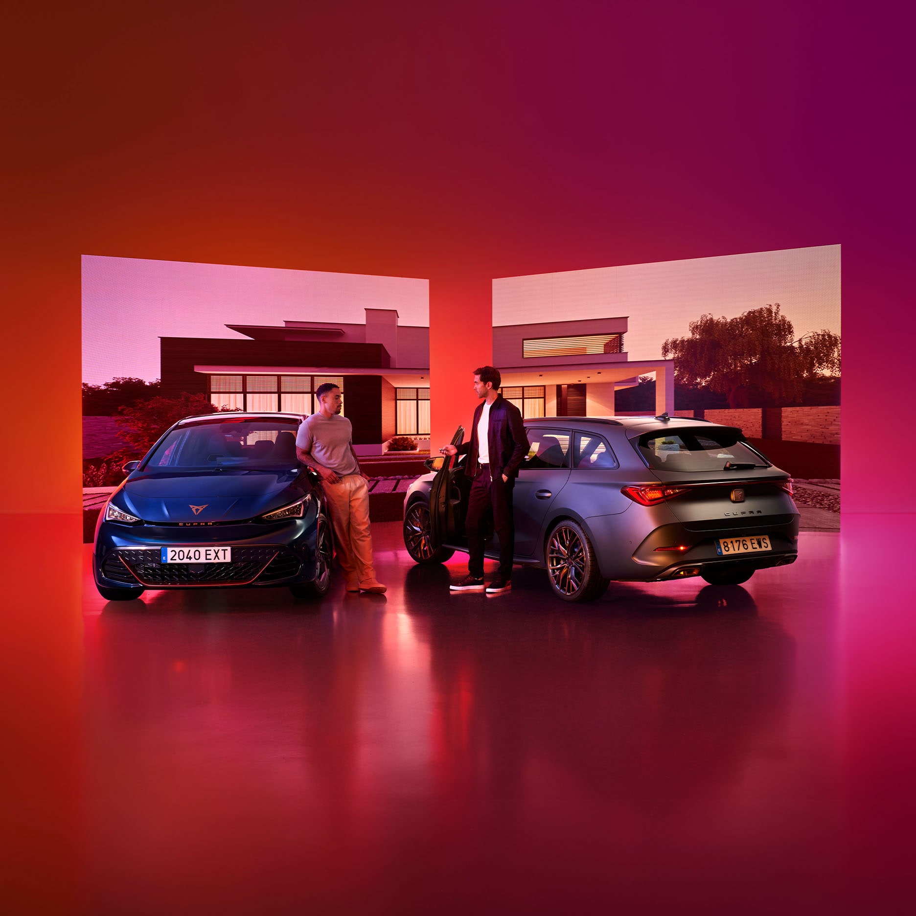 Two CUPRA cars are parked in a studio with a highly reflective floor and warm reddish-purple gradient lighting. Two men stand between the vehicles chatting, framed by two large background screens displaying a modern residential house at dusk. 