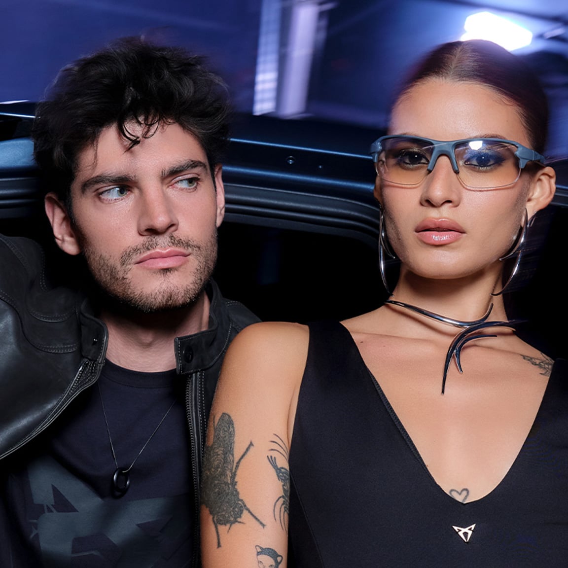 A man in a black leather jacket leans out of a dark CUPRA car door next to a woman wearing sunglasses and large hoop earrings. The woman has a tattoo on her arm and a metallic choker. The scene is dark, lit by interior and ambient blue light.