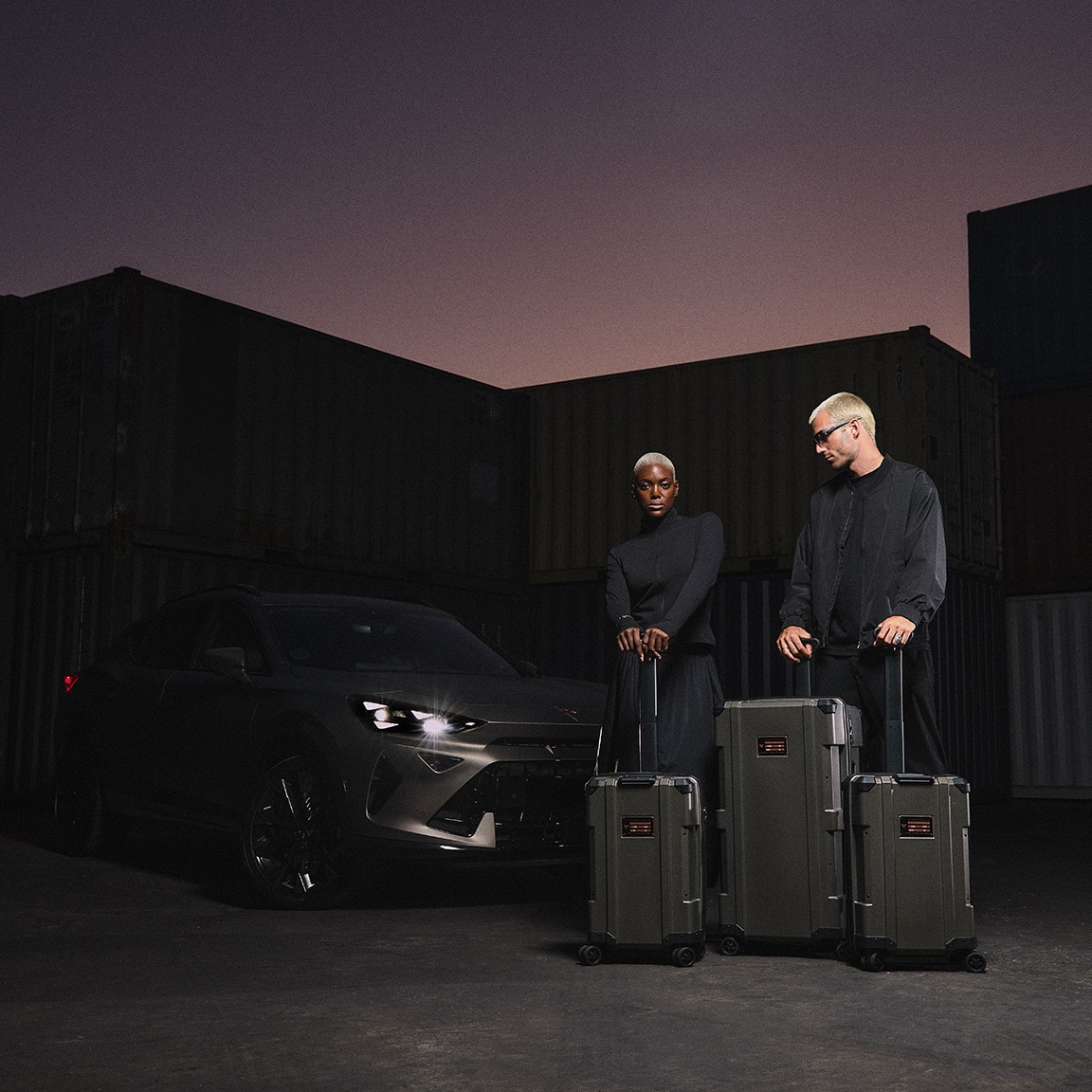A man and a woman, both wearing black, stand with three matching pieces of luggage in front of a dark CUPRA car. The background is a dimly lit industrial container yard at dusk.