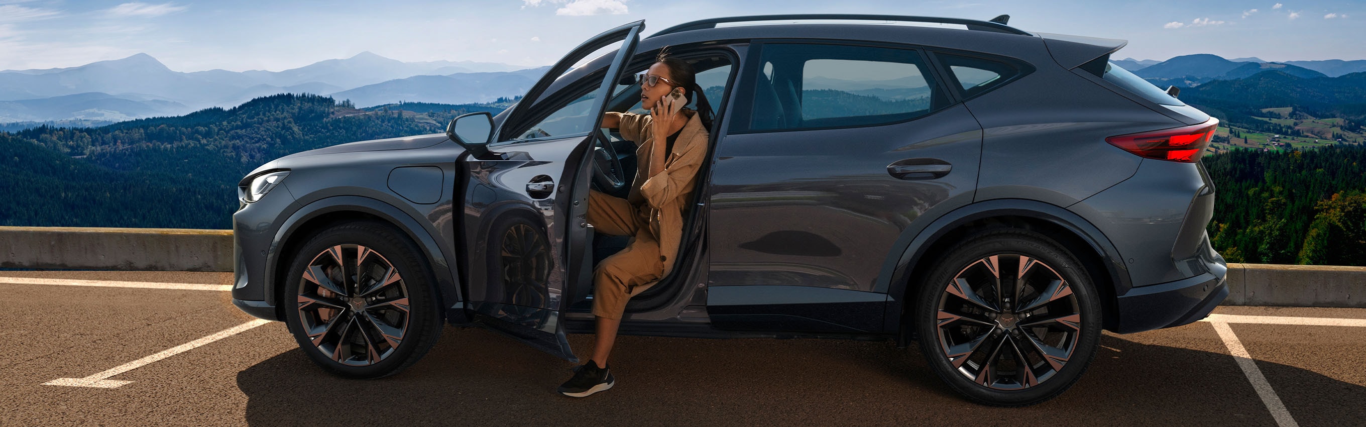 A woman gets out of a dark gray CUPRA car, which is parked in a lot overlooking a scenic mountain range and forested hills. The person is holding a phone to their ear, with the car door open.