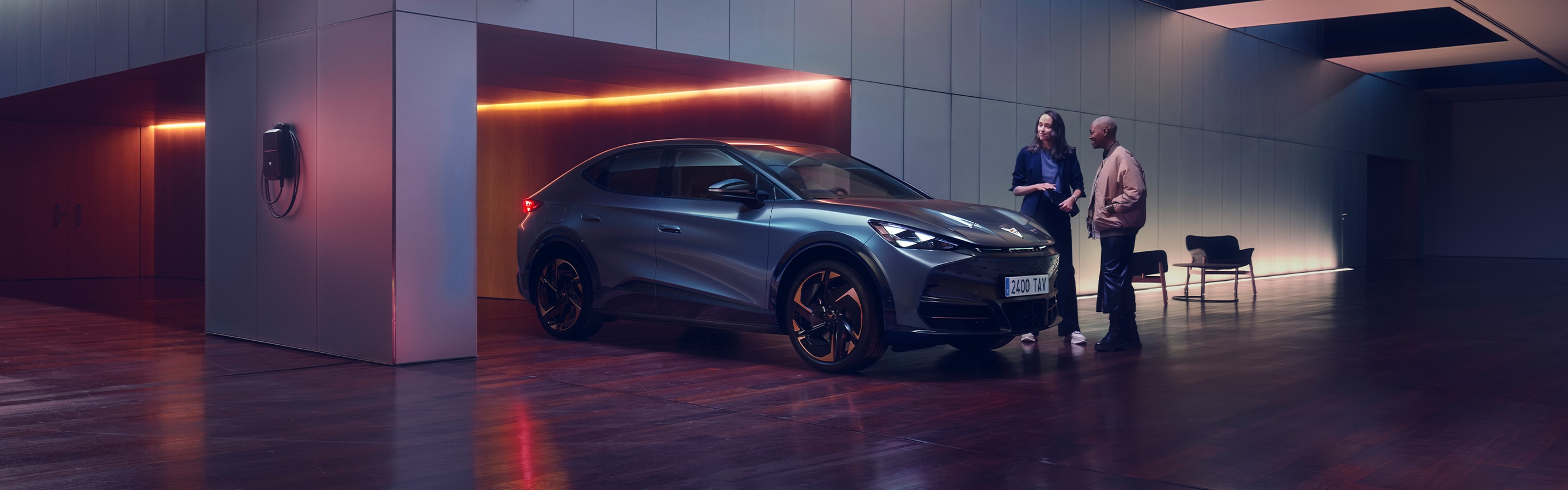  Two people are standing next to a CUPRA car in a sleek, modern, dimly lit showroom with a large skylight. One person is holding a tablet and gesturing towards the car, seemingly explaining something to the other person. A CUPRA logo is mounted on the wall behind them.