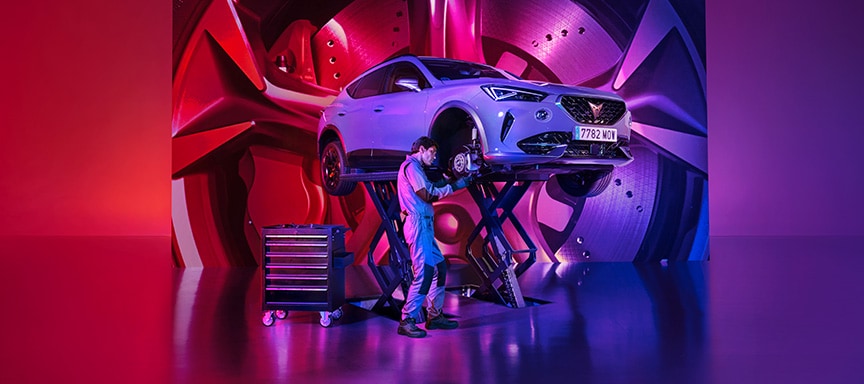 A mechanic works on the front wheel area of a CUPRA car elevated on a scissor lift. The stylized studio setting features a highly reflective floor, a rolling tool cabinet, and a large background screen displaying an oversized, vibrant graphic of a car wheel, all bathed in pink, red, and blue gradient lighting.