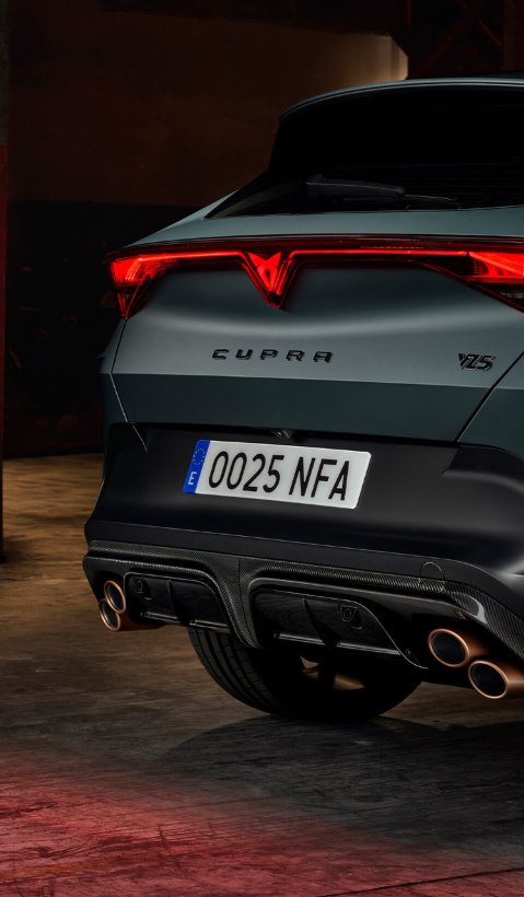 A rear three-quarter view of a CUPRA Formentor parked in a dimly lit garage, showcasing its distinctive illuminated full-width tail light and copper-finished quad exhaust tips.