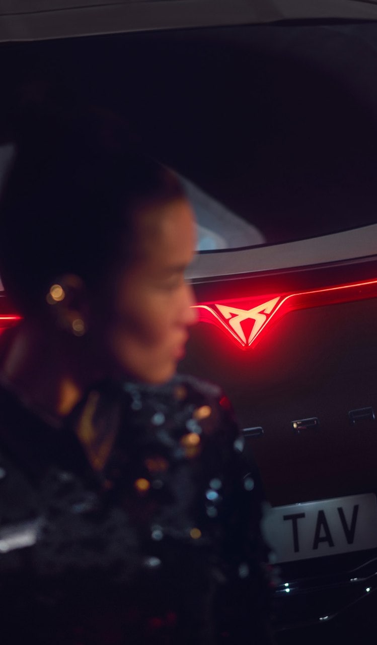 Rear view of a CUPRA Tavascan at night with illuminated red taillights and an integrated glowing logo, featuring a blurred person in the foreground.