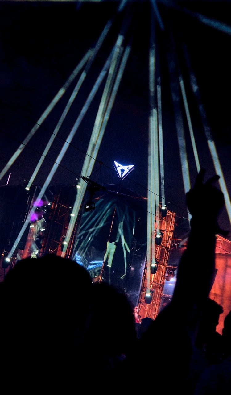 Perspective from a crowd at a night music festival under fanned searchlights from a large stage with a central glowing CUPRA inverted triangle logo.