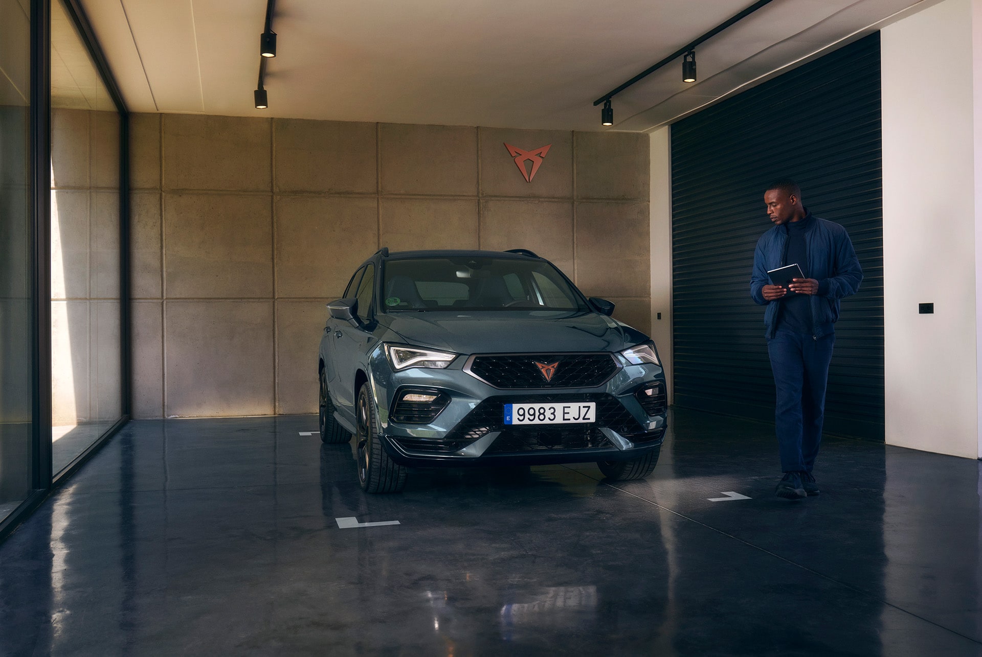 A dark gray CUPRA car is parked inside a modern garage with concrete walls and a polished dark floor. A man is walking toward the car, holding a tablet. A CUPRA logo is mounted on the wall.
