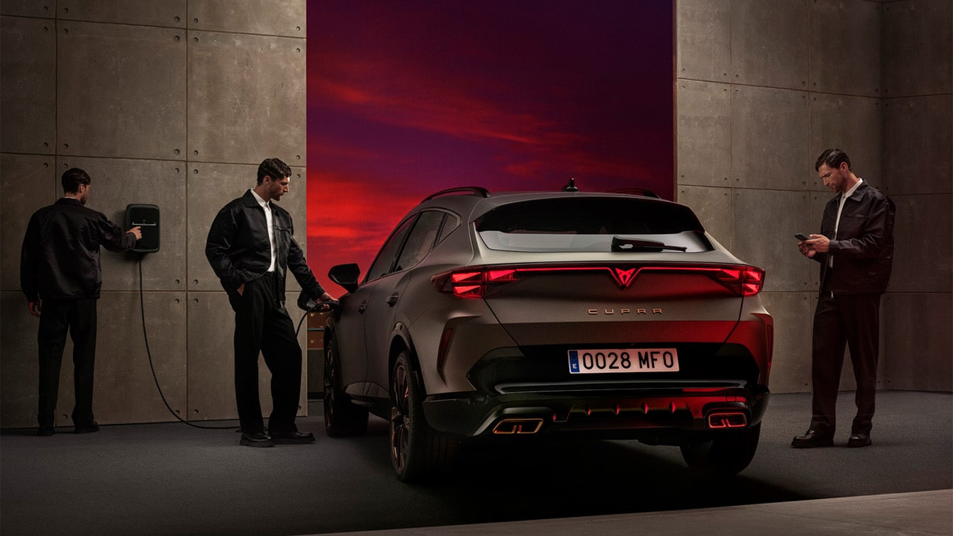 Rear view of a CUPRA car being charged at a wallbox in a modern garage with concrete walls. Three people are present, with one connecting the charging cable and another checking a smartphone, set against a dramatic red sunset sky background.