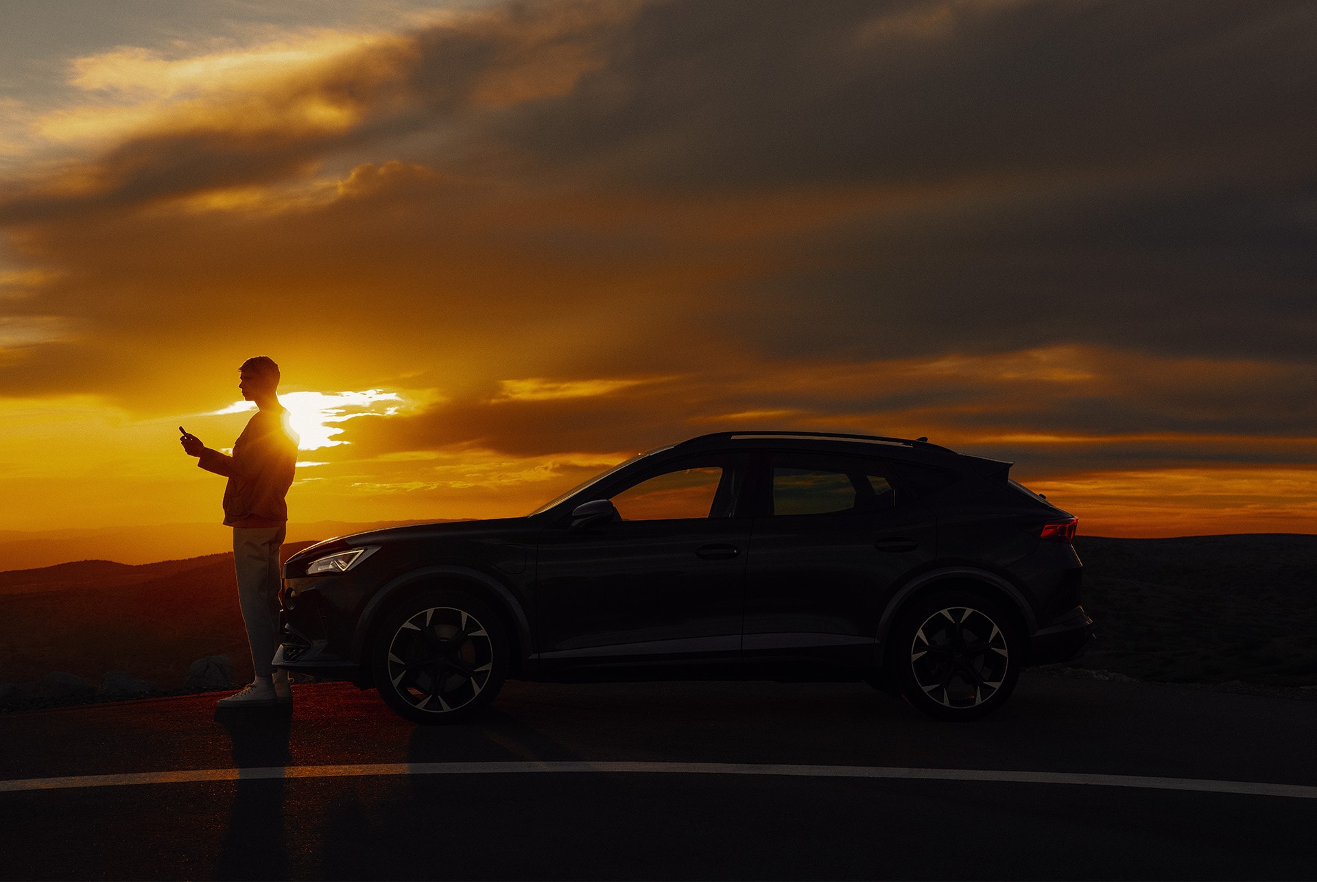 A silhouette of a person standing on the side of a road, looking at a smartphone, next to a dark CUPRA car. The scene is set at sunset, with the sun low on the horizon and dark clouds above.