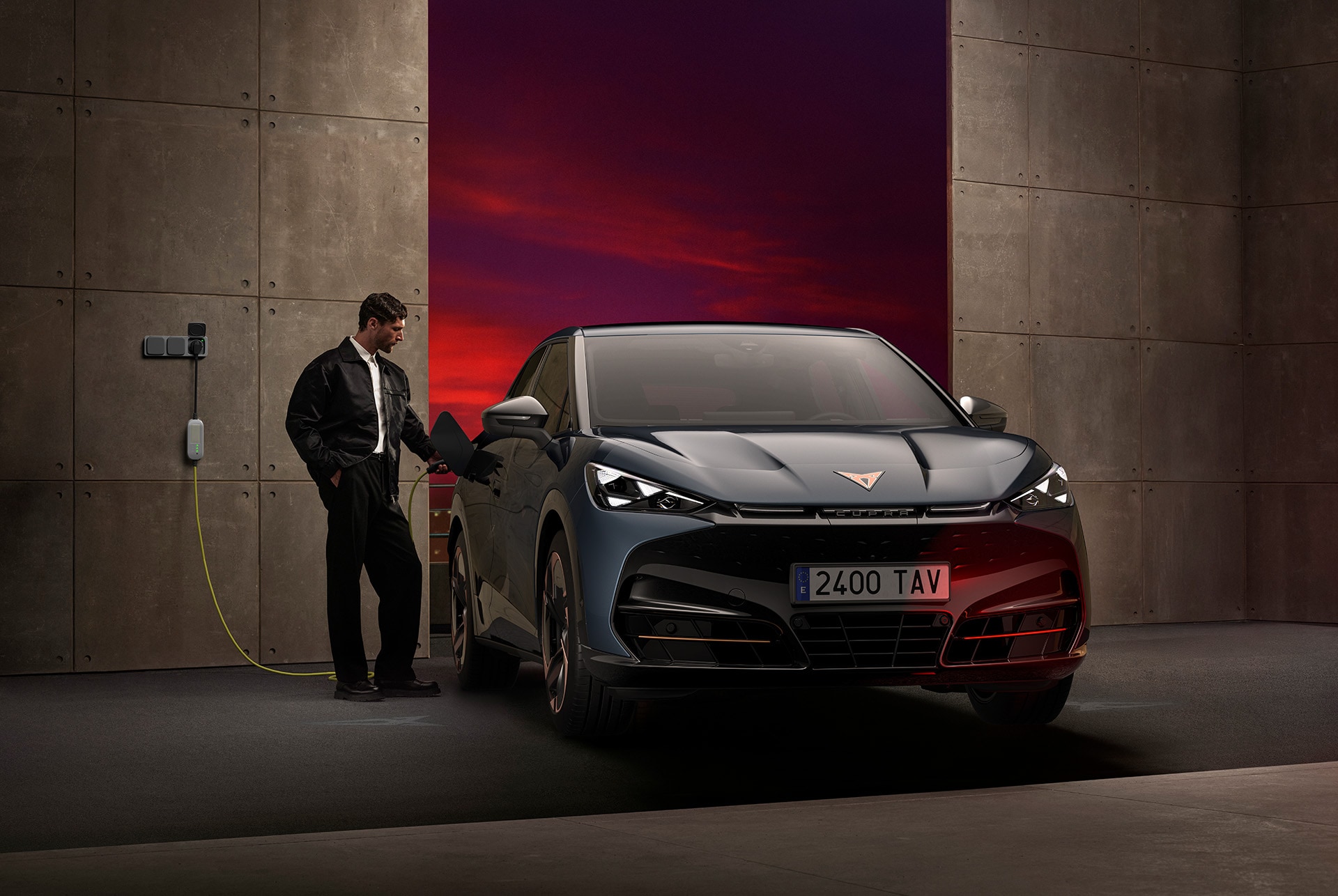 A man is standing next to the front of a dark CUPRA car that is plugged into a wall charger. He is interacting with the car's charging port, and the car is parked in a dark garage with a vibrant red and purple background.
