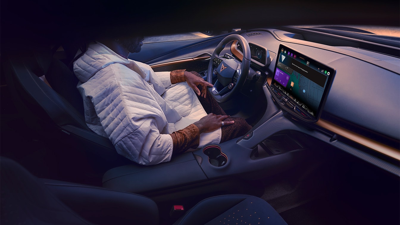Interior view of the CUPRA Tavascan from the passenger side, showing a driver sitting in the front seat with the central infotainment touchscreen displaying the digital assistant interface. 