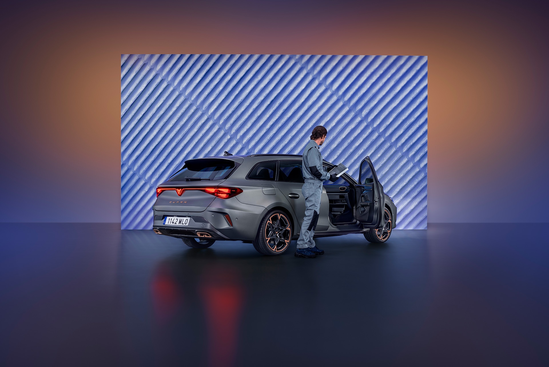 A car technician in a grey jumpsuit stands next to a CUPRA car. The car door is open, and the technician is looking down at a device they are holding. The scene is lit with a blue and orange gradient against a textured backdrop.