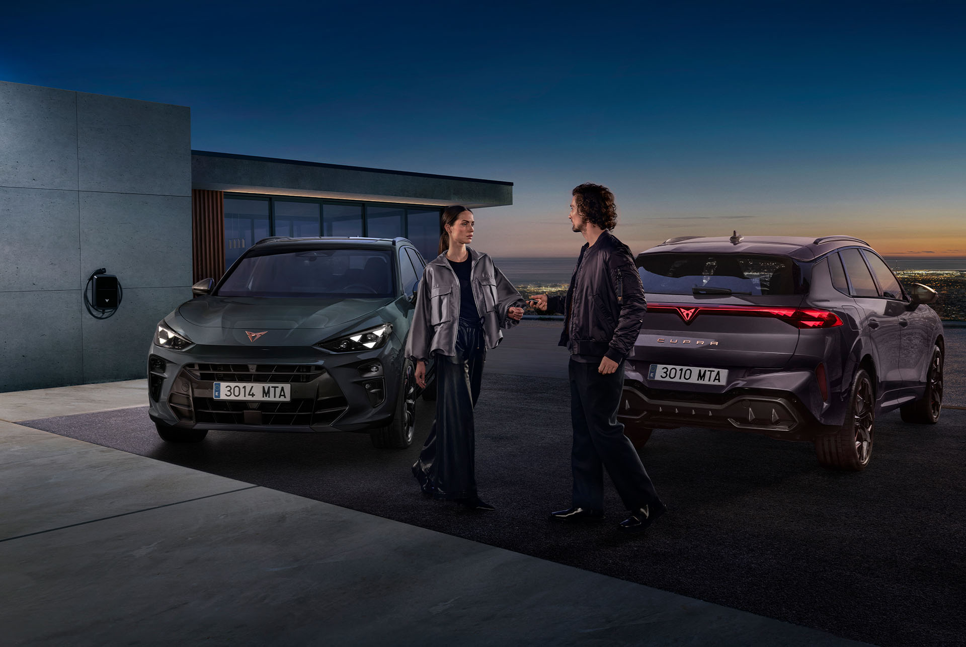 A man and a woman stand between two CUPRA cars parked in the driveway of a modern building at dusk. An electric vehicle charger is visible on the wall, and the sky has a gradient of orange and deep blue.