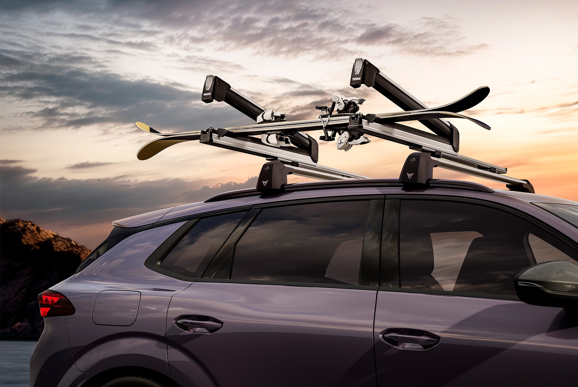 A close-up side view of a dark-colored CUPRA Terramar with two pairs of skis mounted securely on a black roof rack. The background shows a sunset sky.