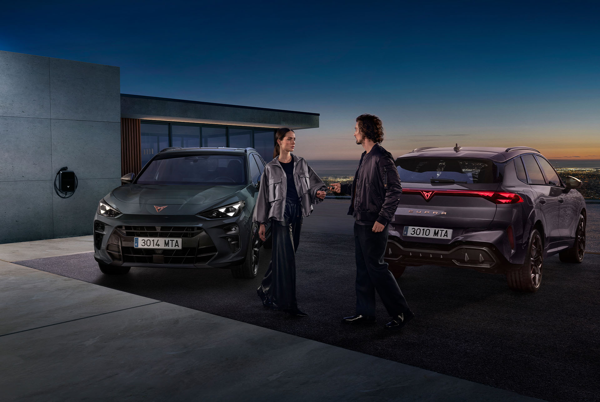 A man and a woman stand between two CUPRA Terramar cars parked in the driveway of a modern building at dusk. An electric vehicle charger is visible on the wall, and the sky has a gradient of orange and deep blue.