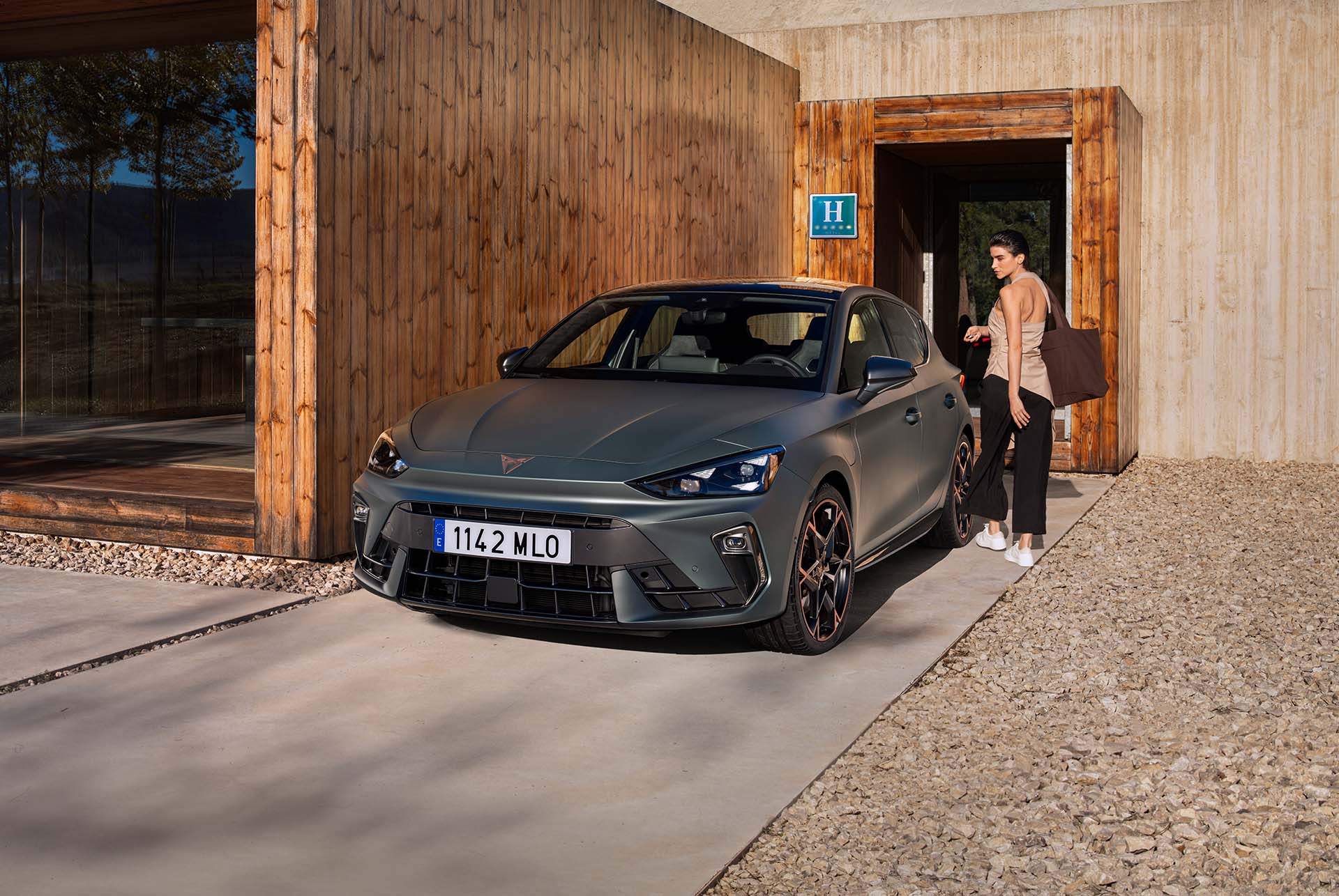 A CUPRA car parked on a concrete driveway in front of a modern house featuring geometric wood paneling and light concrete structures. A woman stands next to the vehicle near a wooden entryway under a bright, partly cloudy sky.