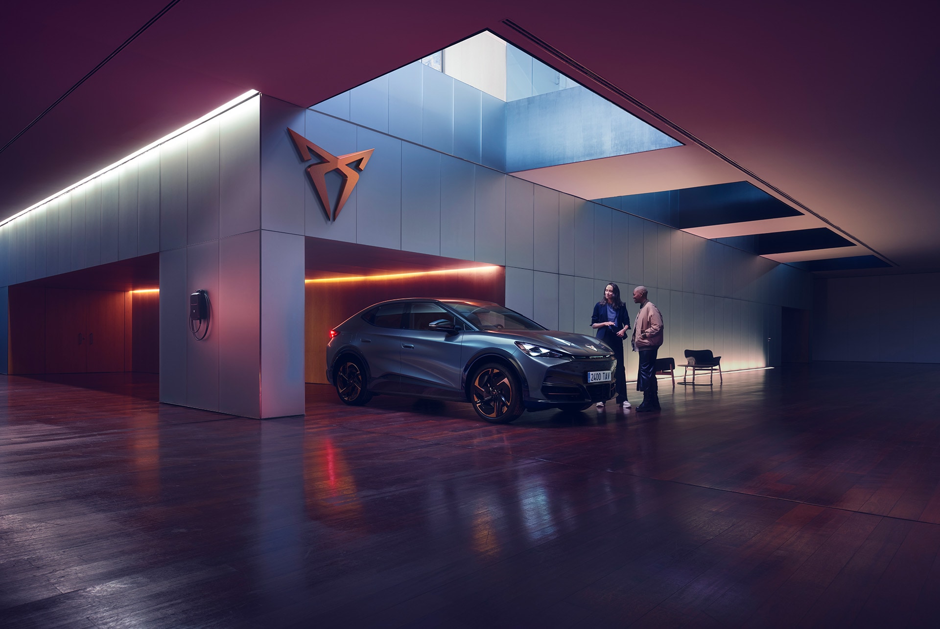 Two people stand next to a CUPRA car in a dark, modern showroom with high-contrast lighting and a large skylight. One person is holding a tablet and talking to the other. A prominent CUPRA logo is mounted on the wall.