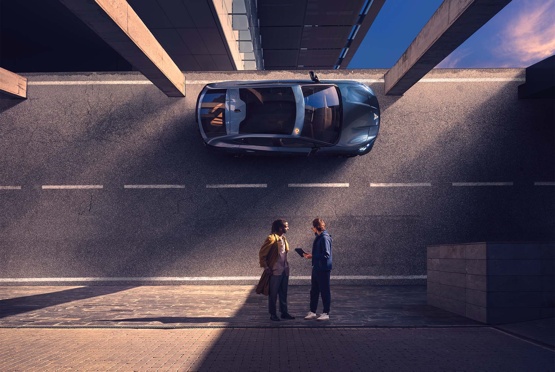 An overhead shot of a dark CUPRA car parked under an overpass or bridge. Two people are standing in front of the car, with one person holding a tablet and speaking to the other. The ground is a mix of asphalt and cobblestone, with strong shadows.