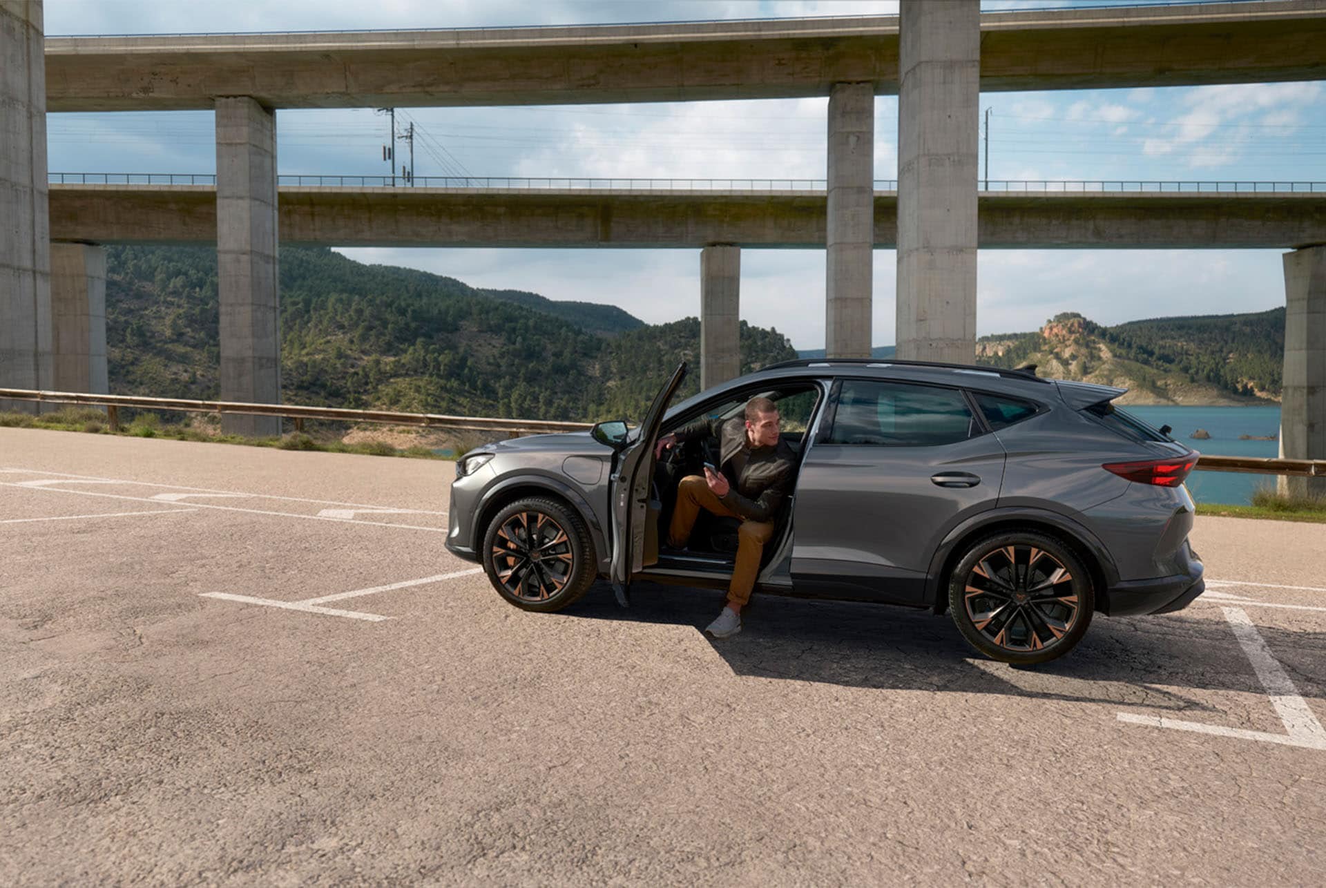A person sits in the driver's seat of a gray CUPRA car with the door open, with their feet on the ground. The car is parked in a lot under a large concrete viaduct, with a lake and mountains in the background.