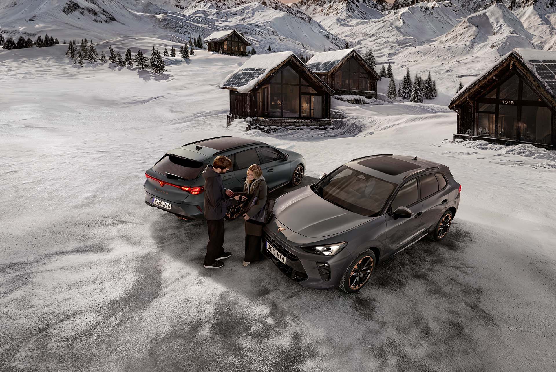 A dark gray CUPRA is parked on a snowy road in a mountain village. A person is standing by the passenger door. In the background, there are several rustic wooden huts and jagged, snow-covered mountains under a sunset sky.