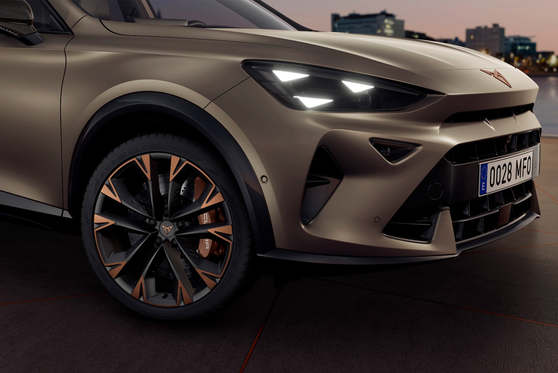 A close-up, angled view of the front of a dark bronze CUPRA car at night. The headlights are on, and the car's distinctive wheel with bronze and black accents is visible. City lights are blurred in the background.