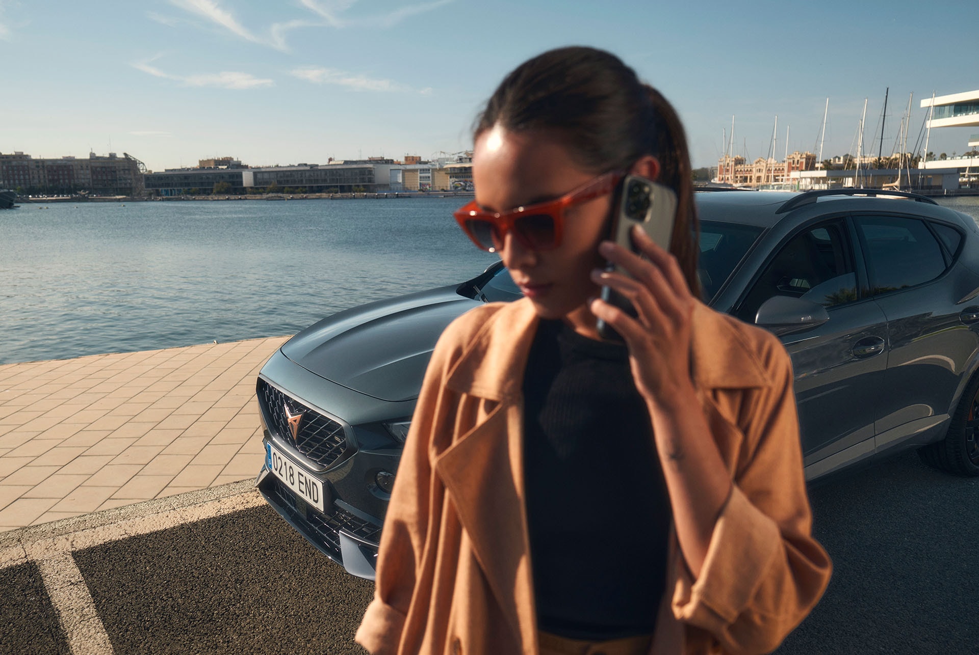 A woman wearing a tan trench coat and sunglasses talking on a smartphone in the foreground, with a CUPRA car parked behind her at a waterfront marina under a clear sky.