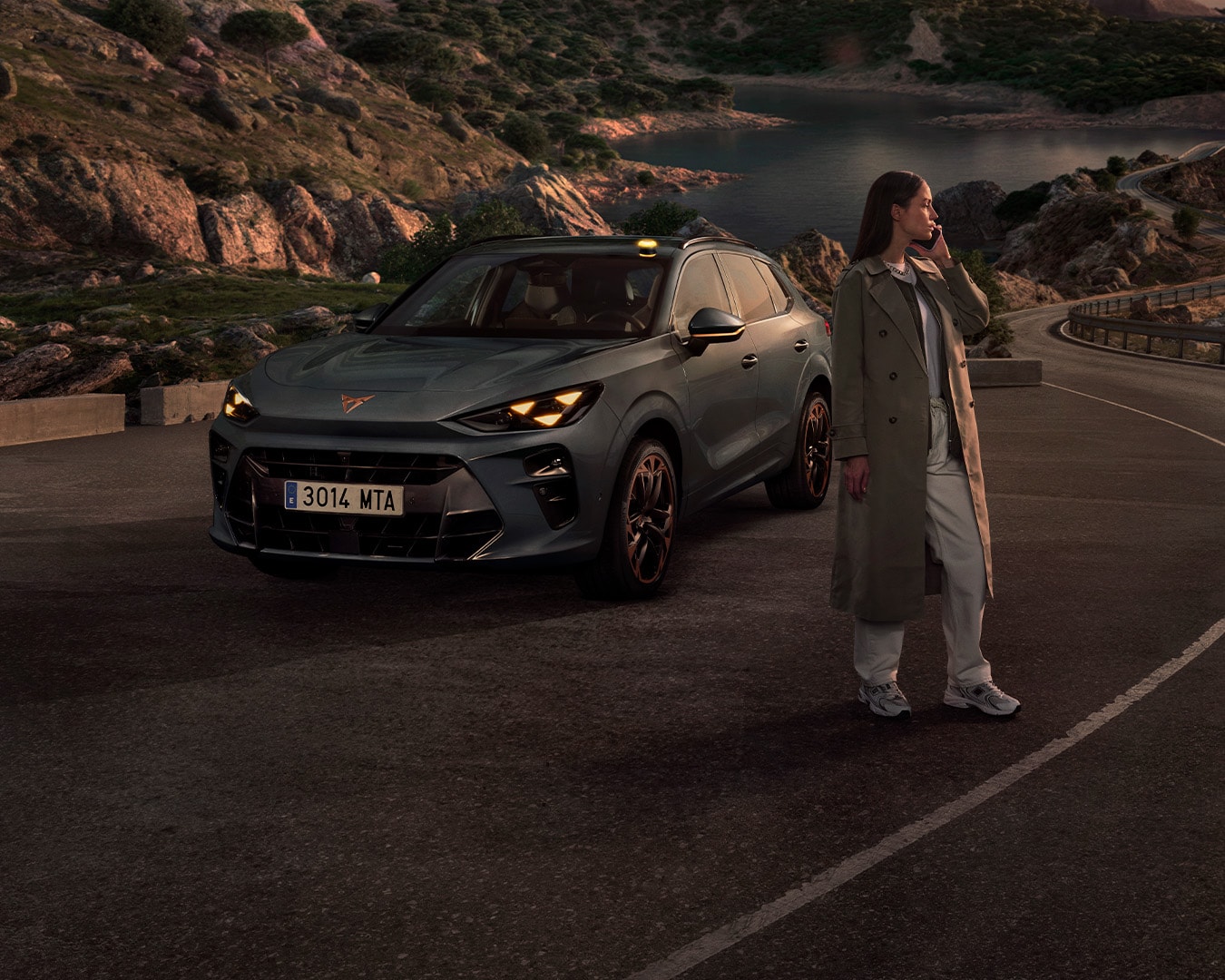A gray CUPRA car is parked on the side of a road at dusk. A person is sitting in the driver's seat with the door open, while two other people stand outside, one on the left and one on the right. The sky has strong orange and blue hues.