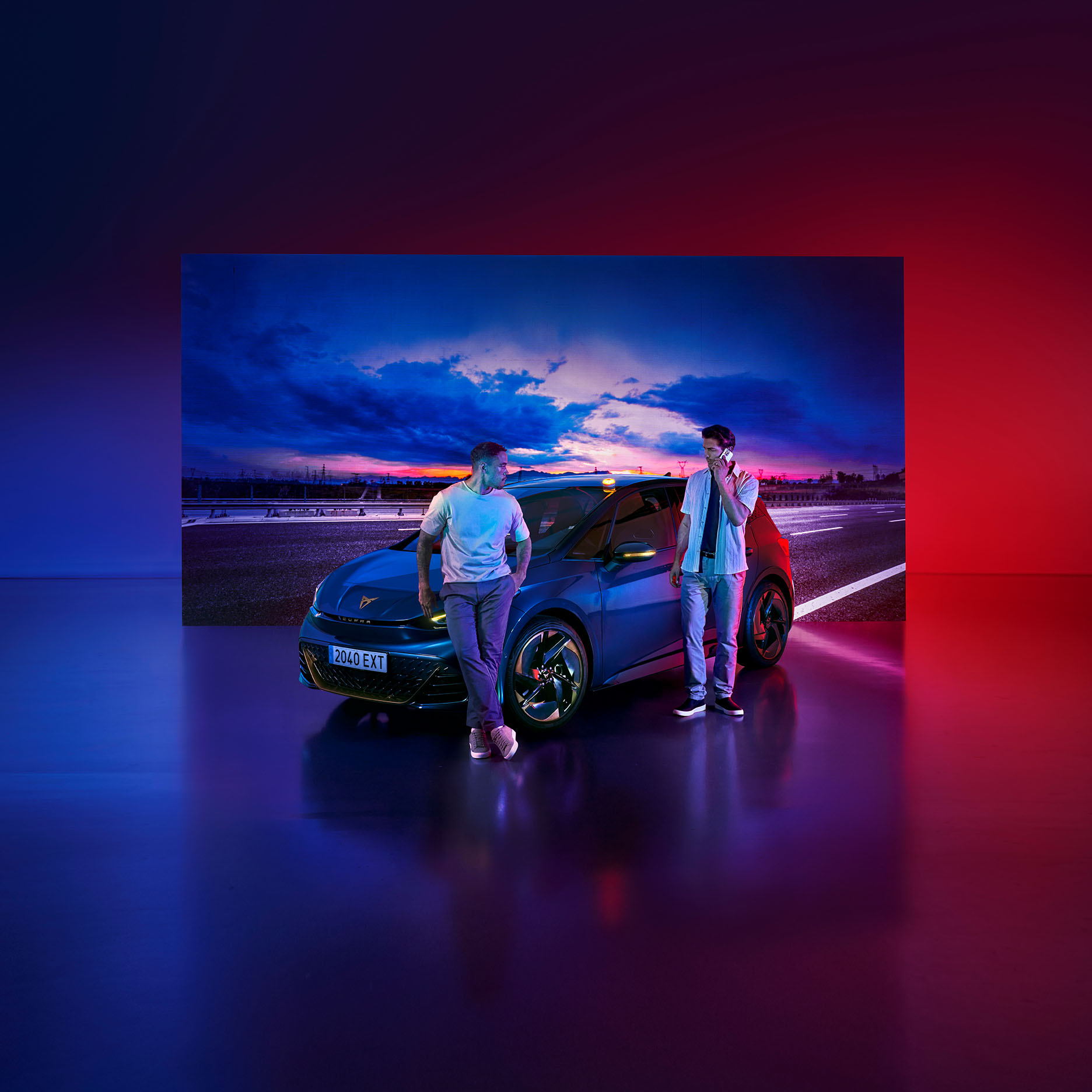 Two people are standing next to a dark blue CUPRA car on an asphalt surface, illuminated by strong red and blue ambient lighting. The background features a twilight sky.