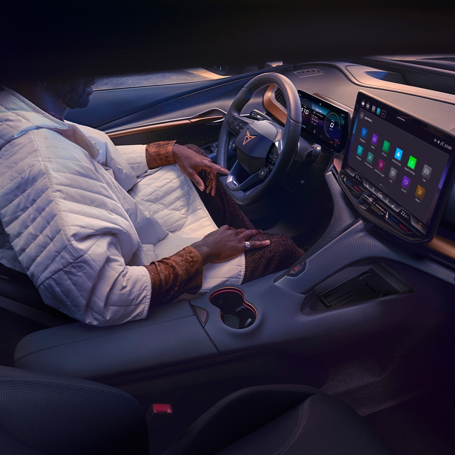 A person wearing a white jacket sits in the driver's seat of a CUPRA car. The view is from the passenger side, showing the dashboard, steering wheel, and a large central touchscreen display.