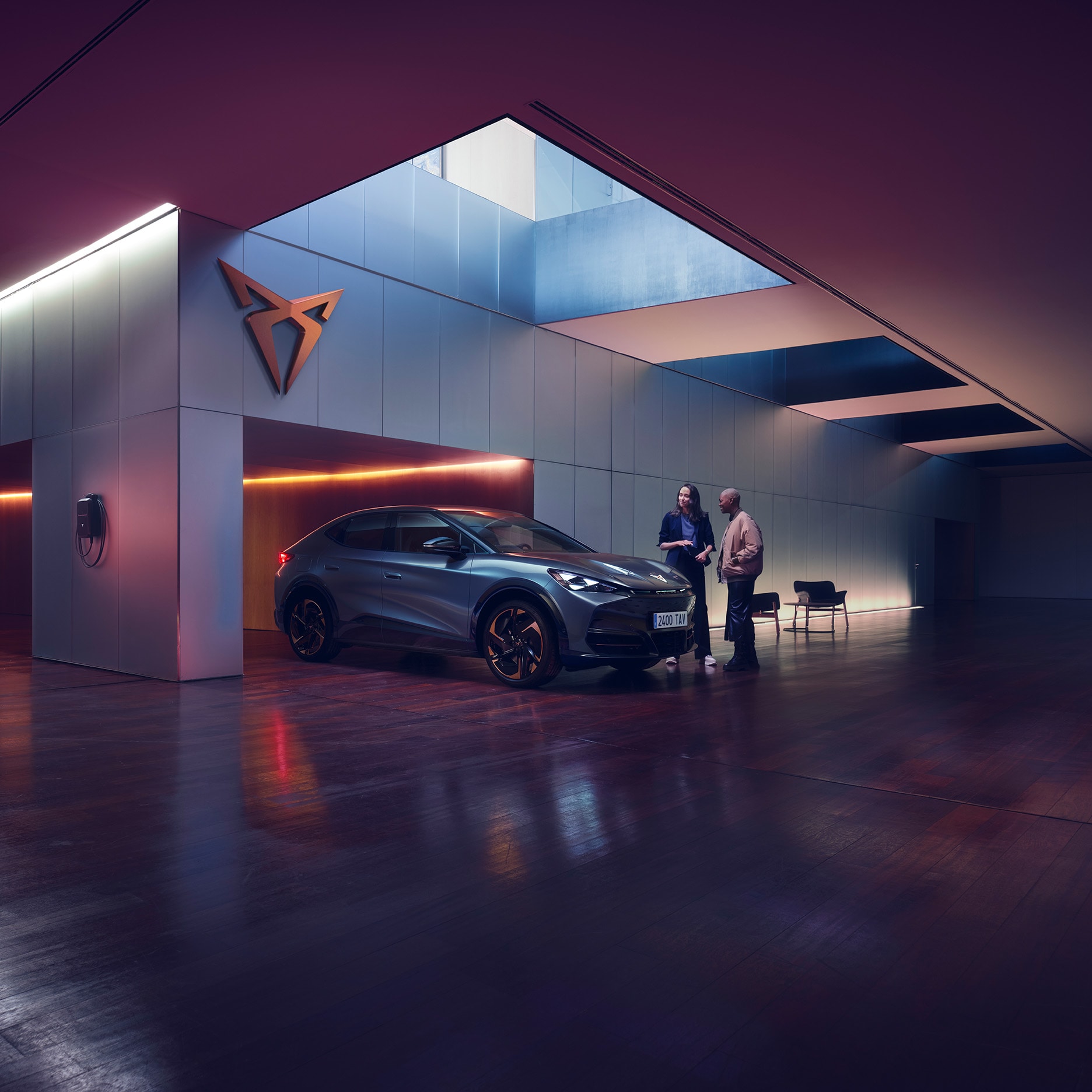 Two people are standing next to a CUPRA car in a sleek, modern, dimly lit showroom with a large skylight. One person is holding a tablet and gesturing towards the car, seemingly explaining something to the other person. A CUPRA logo is mounted on the wall behind them.