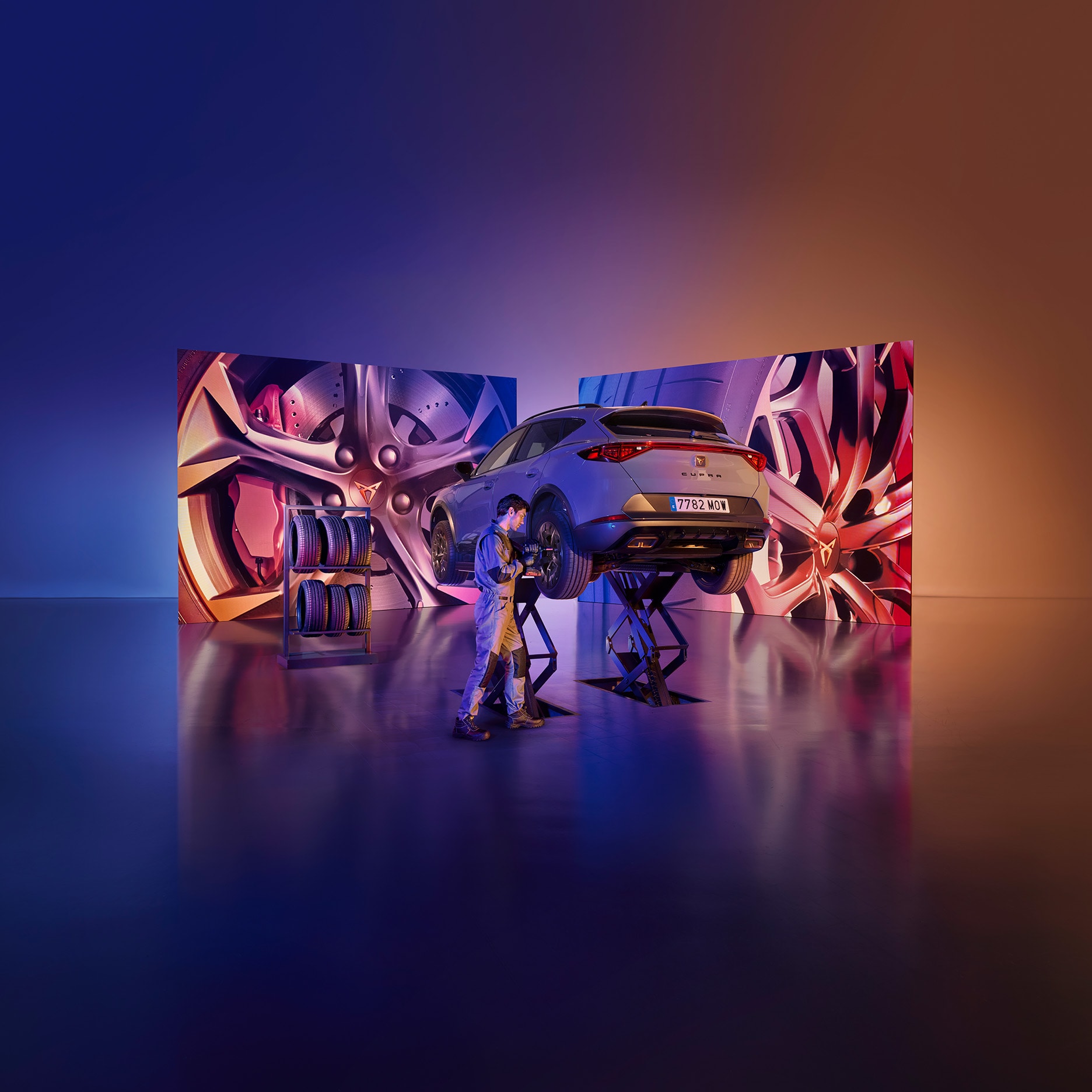 A mechanic is working on a gray CUPRA car that is elevated on a lift in the center of a studio. The background features two large screens with abstract, metallic designs in pink and red. Spare tires are stacked on the side.