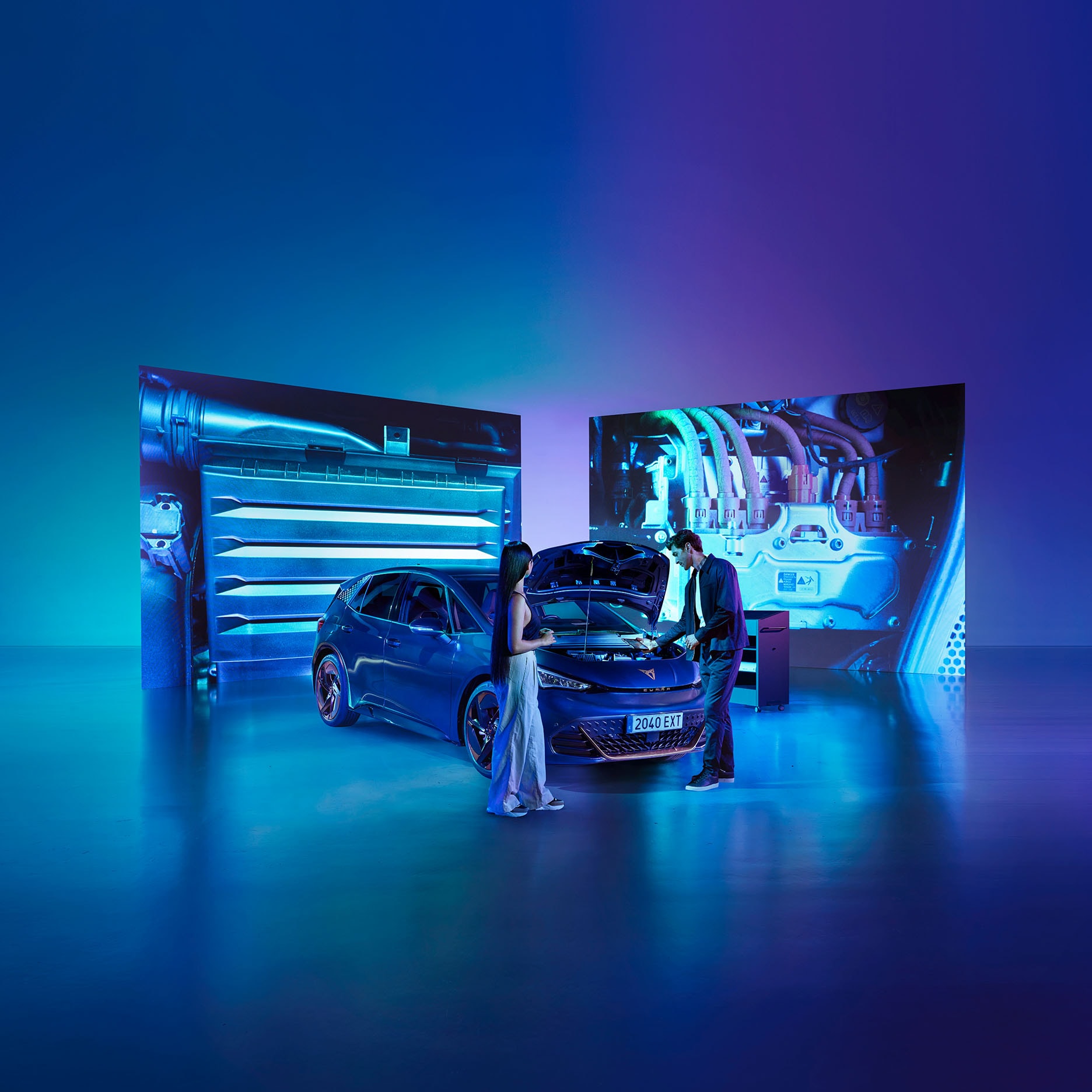 A man and a woman look into the open engine bay of a dark CUPRA car. The scene is set in a futuristic, blue-lit showroom with a reflective floor and large background screens displaying detailed images of the car's internal mechanics.