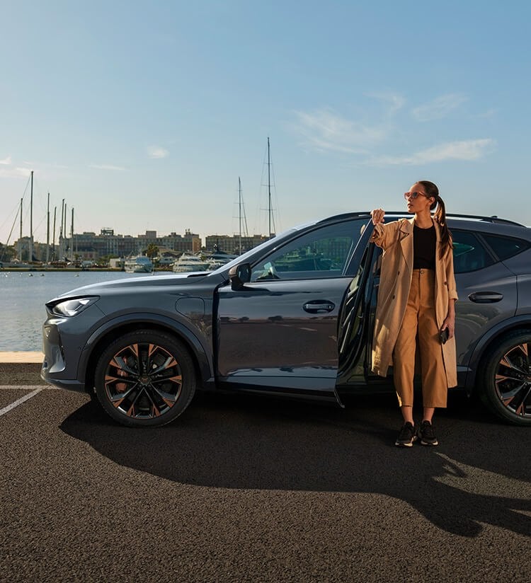 A woman standing beside the open door of a dark grey CUPRA Formentor, parked along a sunny waterfront road, with boats visible in the distance.
